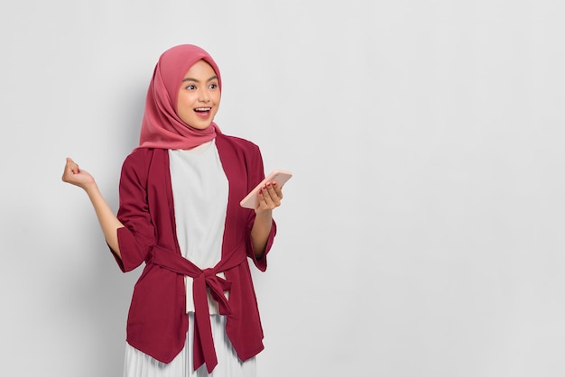 Excitée belle femme asiatique en chemise décontractée et hijab à l'aide d'un téléphone portable, regardant loin l'espace vide isolé sur fond blanc. Concept de mode de vie religieux de personnes