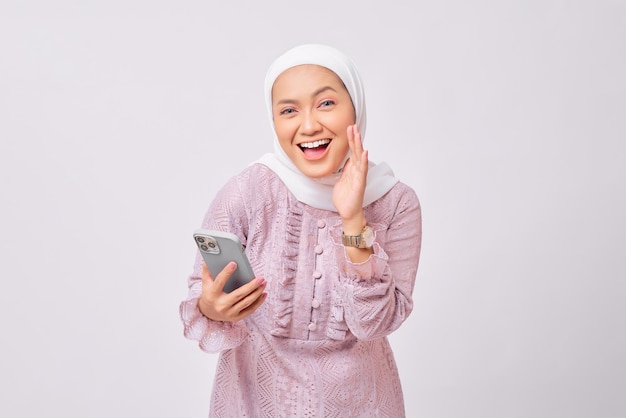 Excitée belle jeune femme musulmane asiatique portant le hijab et la robe violette tenant un téléphone portable et diffusant la promo Ramadan isolée sur fond de studio blanc