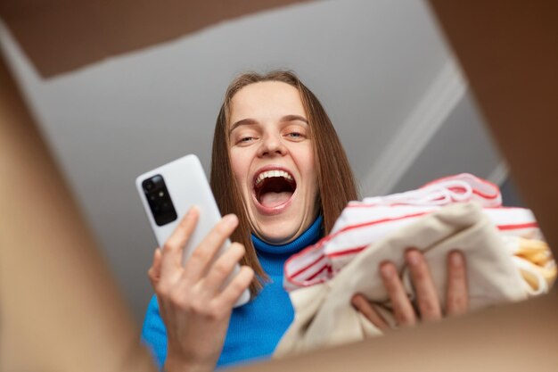 Excitée extrêmement heureuse joyeuse joyeuse femme aux cheveux noirs en chemise bleue regardant à l'intérieur d'une boîte en carton tenant un téléphone intelligent et une pile de vêtements déménageant ou reçu une commande en ligne