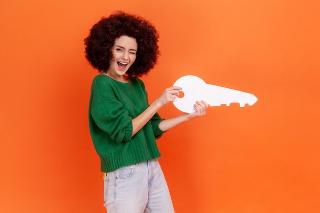 Excitée femme étonnée avec une coiffure Afro portant un chandail vert de style décontracté étant heureuse d'avoir sa propre propriété, tenant une clé en papier dans les mains. Studio intérieur tourné isolé sur fond orange.