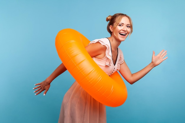 Excitée femme séduisante et heureuse en robe d'été avec des petits pains tenant un anneau en caoutchouc orange et courant vers la piscine, veut se rafraîchir par une chaude journée. Studio intérieur tourné isolé sur fond bleu.