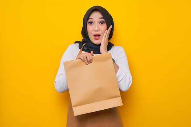 Excitée jeune femme asiatique barista en tablier de t-shirt blanc travail dans un café tenir un sac à emporter en papier vierge maquette isolé sur fond jaune
