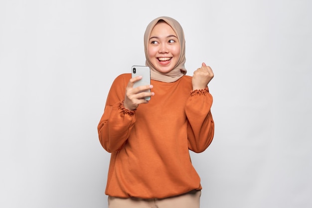 Excitée jeune femme asiatique en chemise orange utilisant un téléphone portable et faisant un geste de gagnant isolé sur fond blanc