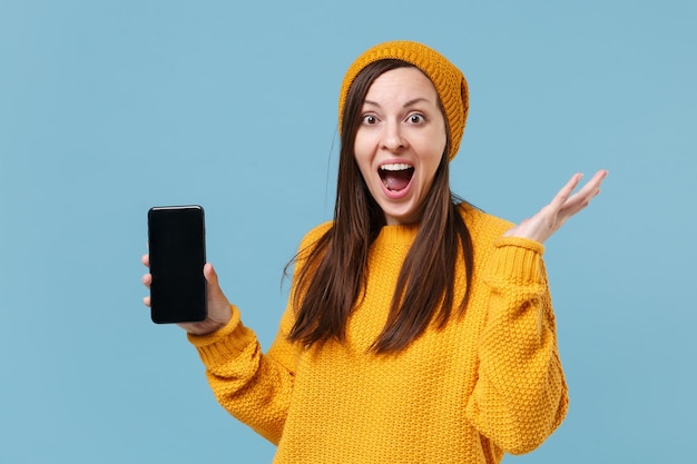 Excitée jeune femme brune fille en pull jaune et chapeau posant isolé sur fond bleu. Concept de style de vie des gens. Maquette de l'espace de copie. Tenez le téléphone portable avec un écran vide vide en écartant les mains.