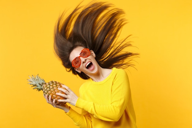 Excitée jeune femme dans des verres amusants avec des cheveux volants tenant des fruits frais d'ananas mûrs isolés sur fond de mur orange jaune. Mode de vie vivant des gens, concept de vacances relaxantes. Maquette de l'espace de copie.