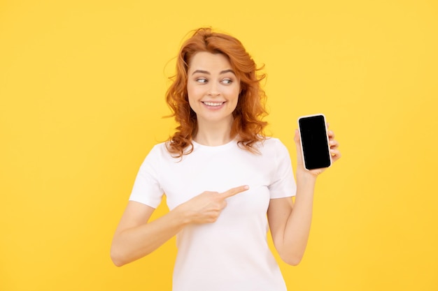 Excitée jeune femme isolée sur fond jaune pointant un téléphone mobile montrant une fille rousse avec un téléphone portable