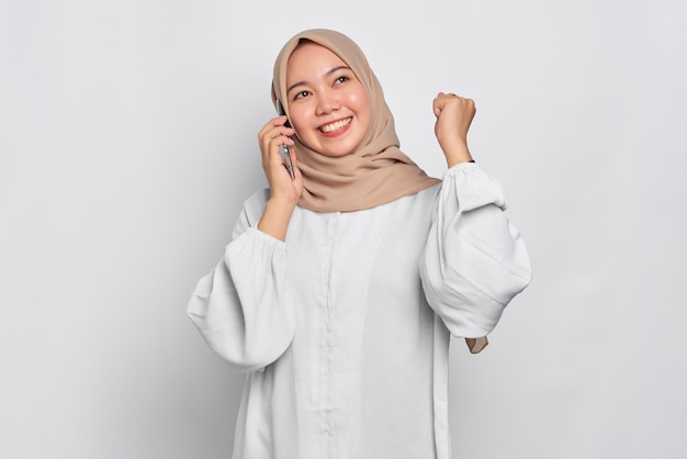 Excitée jeune femme musulmane asiatique parlant au téléphone mobile et faisant le geste du gagnant isolé sur fond blanc