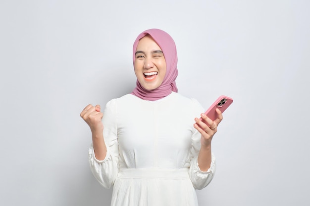 Excitée jeune femme musulmane asiatique utilisant un téléphone portable et célébrant le succès en obtenant de bonnes nouvelles isolées sur fond blanc