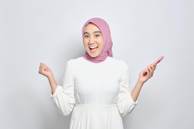 Excitée jeune femme musulmane asiatique utilisant un téléphone portable et célébrant le succès en obtenant de bonnes nouvelles isolées sur fond blanc