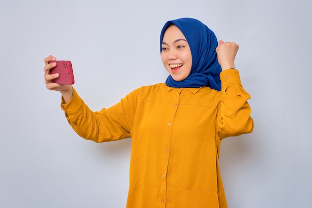Excitée jeune femme musulmane asiatique vêtue d'une chemise orange jouant à un jeu en ligne sur un téléphone mobile célébrant la victoire isolée sur fond blanc