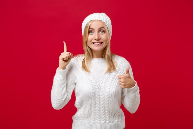 Photo excitée jeune femme en pull blanc, chapeau isolé sur fond rouge. mode de vie sain, concept de saison froide. maquette de l'espace de copie. tenir l'index vers le haut avec une excellente nouvelle idée, montrant le pouce vers le haut.