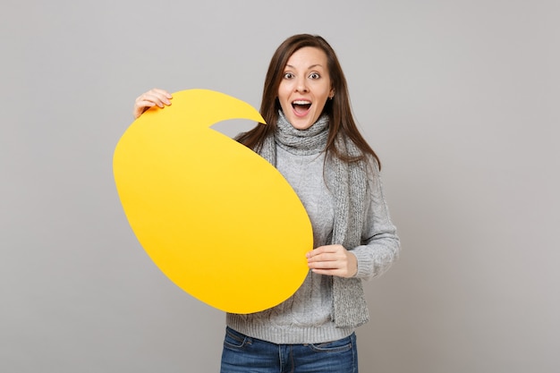 Excitée jeune femme en pull gris, écharpe tenant un blanc vide jaune Dites bulle de dialogue nuage isolée sur fond de mur gris. Mode de vie sain, émotions sincères, concept de saison froide.