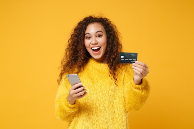 Excitée jeune fille afro-américaine en pull de fourrure posant isolé sur fond de mur orange jaune en studio. Concept de style de vie des gens. Maquette de l'espace de copie. Tenir un téléphone portable, une carte bancaire de crédit.