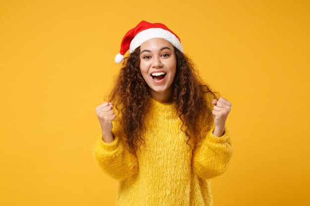 Excitée jeune fille afro-américaine Santa en pull de fourrure, chapeau de Noël isolé sur fond jaune. Bonne année 2020 concept de vacances de célébration. Maquette de l'espace de copie. Serrer les poings comme vainqueur.