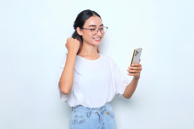 Excitée jeune fille asiatique en t-shirt blanc à l'aide d'un smartphone et faisant un geste gagnant isolé sur fond blanc