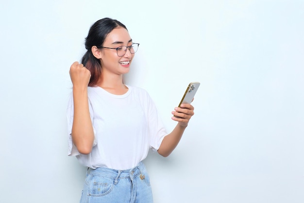 Excitée jeune fille asiatique en t-shirt blanc à l'aide d'un smartphone et faisant un geste gagnant isolé sur fond blanc