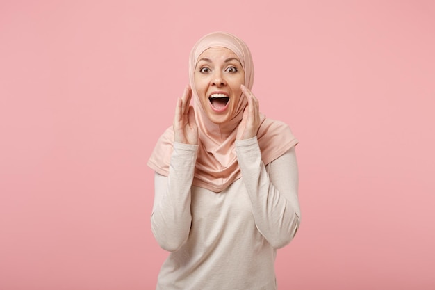 Excitée jeune fille musulmane arabe en vêtements légers hijab posant isolé sur fond rose en studio. Concept de style de vie de l'islam religieux des gens. Maquette de l'espace de copie. Crier avec les mains près de la bouche.