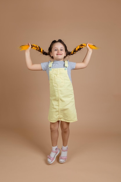 Excitée jeune fille tenant des tresses kanekalon jaunes sur le point de sauter en regardant la caméra en souriant portant une combinaison jaune et un t-shirt gris sur fond beige