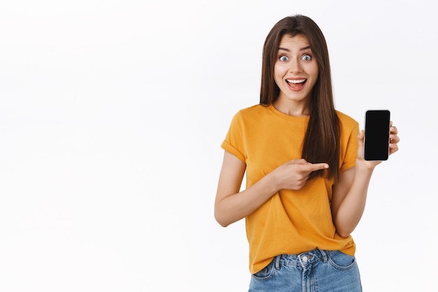 Excitée ravie jeune jolie femme en t-shirt jaune montrant l'écran du smartphone fixement étonné et amusé souriant fasciné pointant l'écran mobile trouvé une application incroyable fond blanc