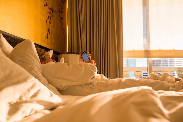 Exécutif de femme adulte allongé sur le lit de l'hôtel au soleil tôt le matin en regardant un téléphone intelligent