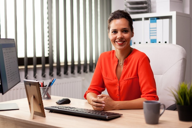 Exécutif indépendant finance entrepreneurur portant un chemisier rouge souriant à la caméra sur le lieu de travail. Femme confiante réussie dans le marketing assise au bureau sur le lieu de travail à l'aide d'un ordinateur.