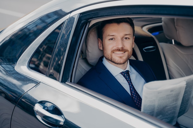 Exécutif professionnel jeune gestionnaire masculin barbu en costume élégant lisant joyeusement des articles de rapport financier dans le journal du matin tout en étant assis pendant le trajet pour travailler dans une voiture de luxe, souriant à la caméra