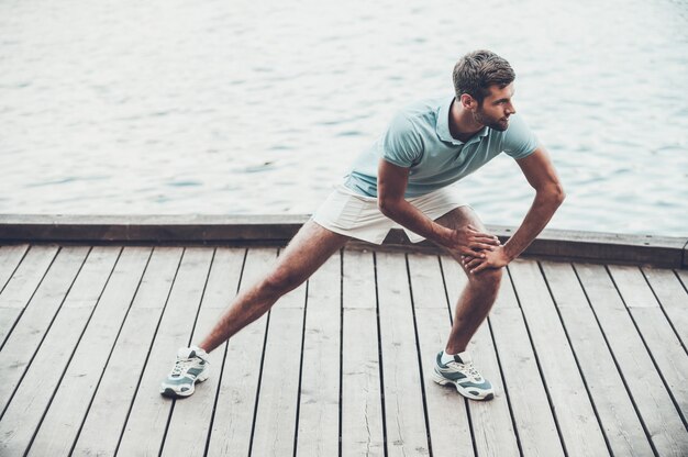 Des exercices d'étirement. Jeune homme confiant faisant des exercices d'étirement en se tenant debout sur le quai