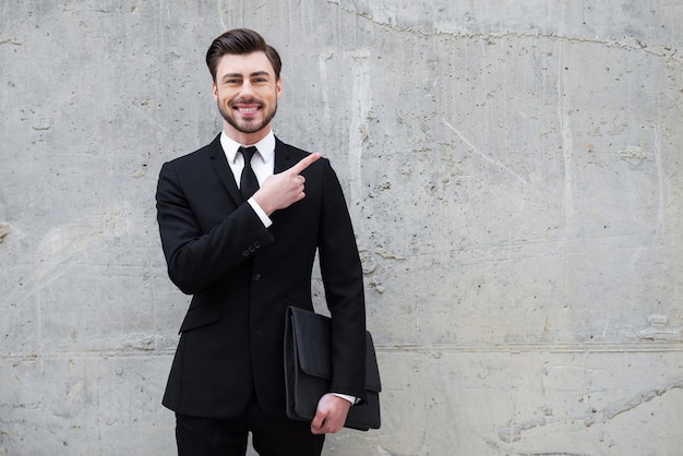 Expert fiable. Beau jeune homme tenant une mallette en se tenant debout à l'extérieur et contre le mur de béton