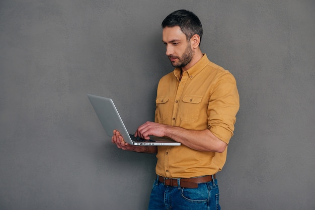 Expert en informatique confiant. Homme mûr confiant travaillant sur un ordinateur portable en se tenant debout sur fond gris