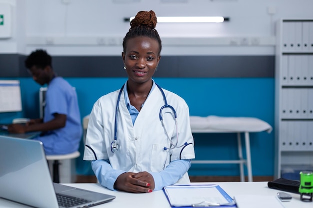 Expert spécialiste de la santé généraliste assis au bureau dans l'armoire de la clinique. Spécialiste polyclinique afro-américain portant un manteau blanc et un stéthoscope assis au bureau.