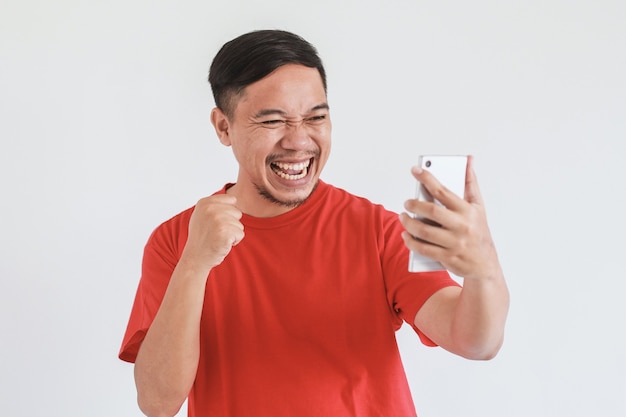 Expression du visage souriant d'un homme asiatique heureux portant un t-shirt rouge regardant le téléphone intelligent avec la main de poing