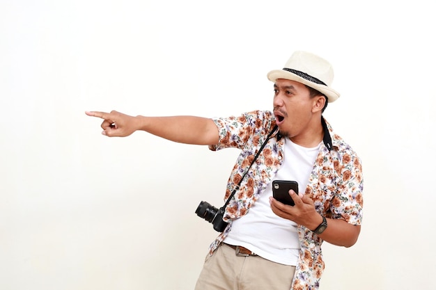 Photo l'expression wow d'un homme adulte asiatique touriste pointant de côté tout en tenant un téléphone portable concept de voyage isolé sur fond blanc