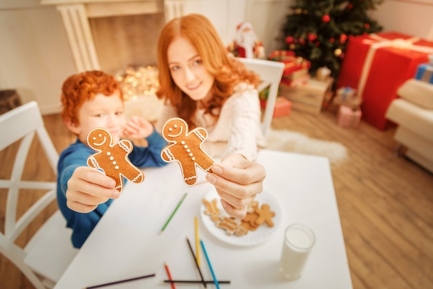 Extrêmement délicieux. Mise au point sélective sur une paire de biscuits en pain d'épice tenus par une mère joyeuse et son petit fils assis à une table et dessinant.