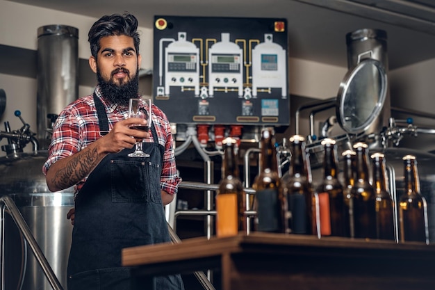 Fabricant masculin hipster tatoué barbu indien dégustant et présentant de la bière artisanale dans la microbrasserie.