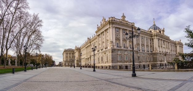 Façade latérale du palais royal de Madrid, rue piétonne avec lampadaires, arbres et journée ensoleillée avec nuages. Espagne.