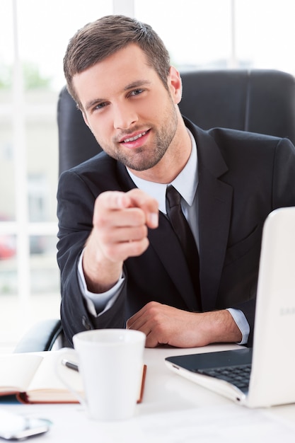 Faciliter les affaires. Jeune homme confiant en tenue de soirée vous pointant et souriant alors qu'il était assis sur son lieu de travail