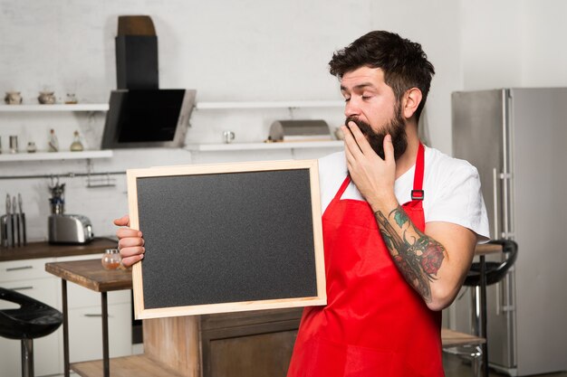 Des façons intelligentes d'organiser la cuisine. Cook tenir l'espace de copie de tableau vierge. Conseils secrets. Informations utiles. Tablier rouge hipster barbu homme stand dans la cuisine. Magasin de meubles de cuisine. Concept de hacks de cuisine.