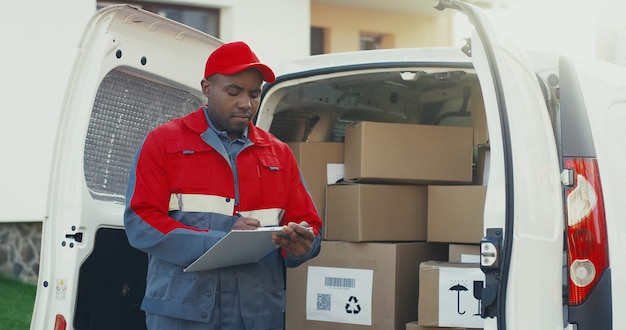 Facteur afro-américain en uniforme rouge et capuchon debout à la camionnette blanche avec des boîtes en carton et en remplissant les documents sur le presse-papiers. Extérieur.