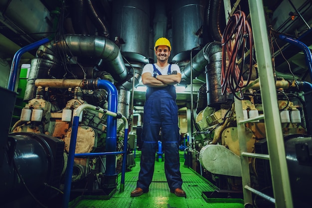 Faible angle de vue du beau travailleur caucasien dans l'ensemble et casque sur la tête debout dans le navire à côté du moteur avec les bras croisés.