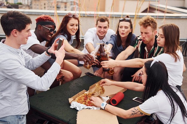 Faire des acclamations par des bouteilles avec de la bière Un groupe de jeunes en tenue décontractée fait la fête ensemble sur le toit pendant la journée