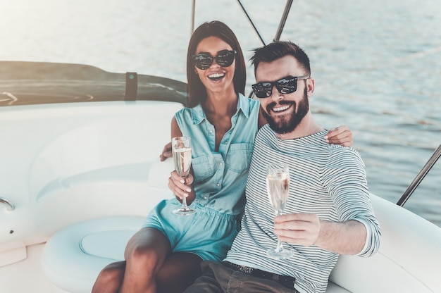 Faire de beaux souvenirs ensemble. Joyeux jeune couple tenant des verres de champagne et regardant la caméra alors qu'il était assis sur le bord du yacht