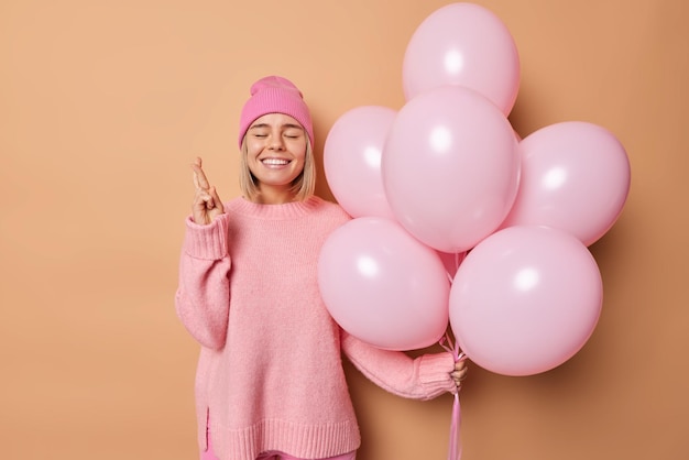 Faire le concept de souhait. Une fille d'anniversaire positive croise les doigts croit que le miracle se produit tient un tas de ballons gonflés porte un chapeau et un pull décontracté isolé sur fond beige célèbre l'événement