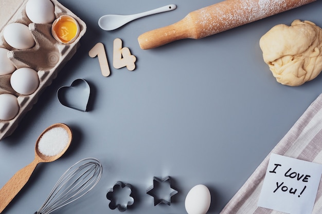 Faire des cookies pour la Saint-Valentin, mise à plat, vue de dessus, espace de copie. Divers ingrédients, rouleau à pâtisserie, pâte, œufs, numéros 14, fouet sur fond gris, mise en page sur fond gris.