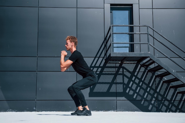 Faire de l'exercice près d'un bâtiment avec un mur noir Jeune homme sportif en chemise et pantalon noirs à l'extérieur pendant la journée