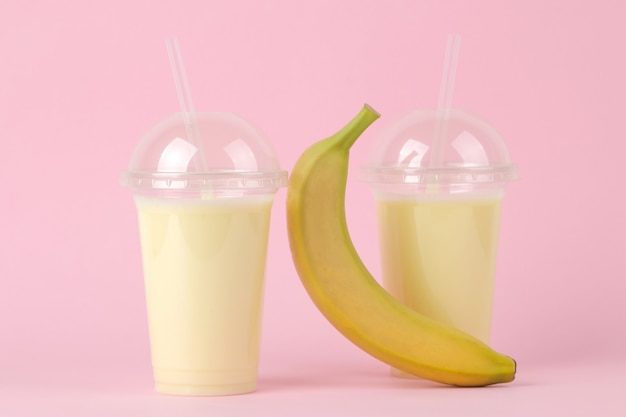 Faire un milk-shake. un verre jetable en plastique avec un milk-shake à la banane et une banane fraîche sur une table rose tendance et lumineuse.