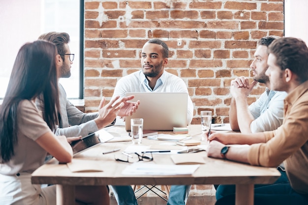 Faire une nouvelle stratégie. Groupe de jeunes discutant de quelque chose assis à la table en bois du bureau