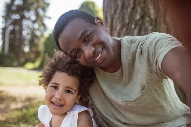 Faire une photo. Homme souriant à la peau sombre faisant selfie avec sa petite fille dans le parc