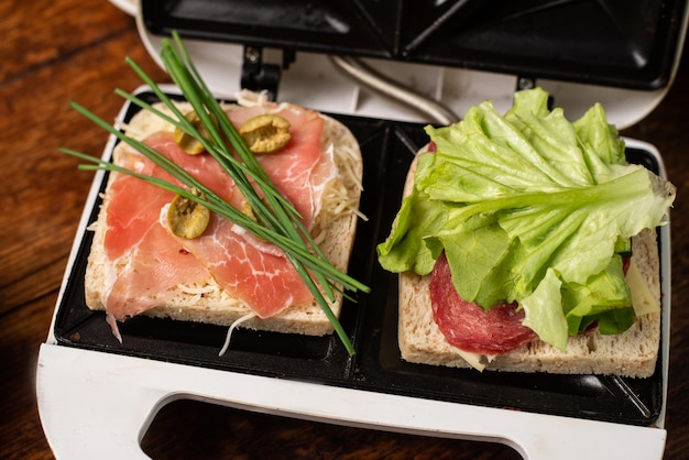Faire des sandwichs à partir de jambon, de légumes et de fromage dans une machine à sandwich Deux toasts avec différentes garnitures en gros plan