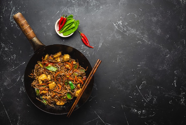 Faire sauter des nouilles soba avec du poulet, des légumes dans une vieille poêle à wok rustique, des baguettes sur fond de pierre noire, gros plan, vue de dessus. Repas traditionnel asiatique/thaï, espace pour le texte