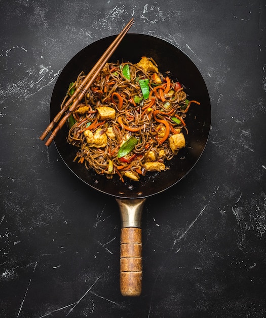 Faire sauter des nouilles soba avec du poulet, des légumes dans une vieille poêle à wok rustique, des baguettes sur fond de pierre noire, gros plan, vue de dessus. Repas traditionnel asiatique/thaï, gros plan